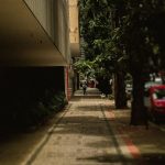 Calçada arborizada em uma rua urbana, com árvores, carro e edifícios ao fundo.