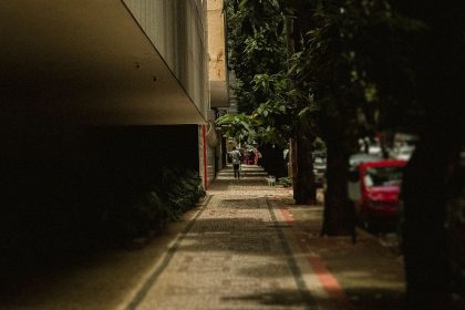 Calçada arborizada em uma rua urbana, com árvores, carro e edifícios ao fundo.