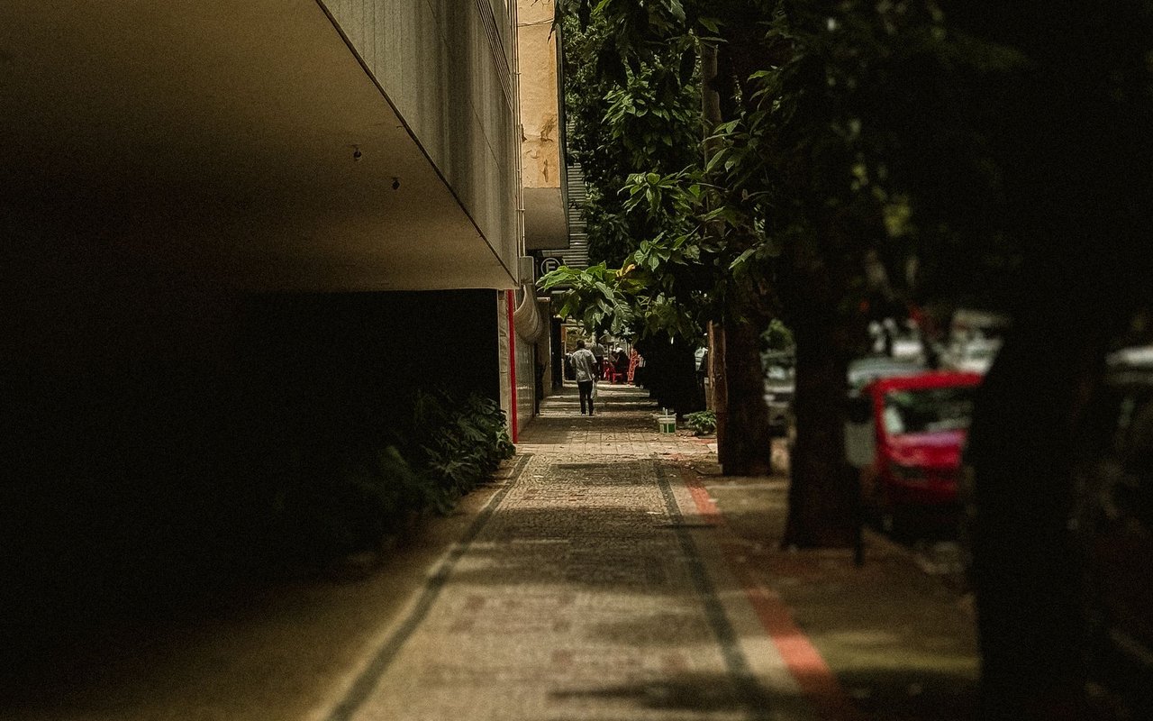 Calçada arborizada em uma rua urbana, com árvores, carro e edifícios ao fundo.