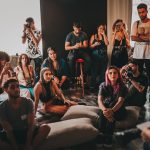 Grupo diverso de pessoas reunido em um espaço amplo, ouvindo atentamente uma mulher que fala sentada no centro.