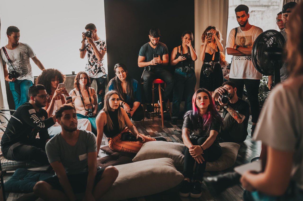 Grupo diverso de pessoas reunido em um espaço amplo, ouvindo atentamente uma mulher que fala sentada no centro.