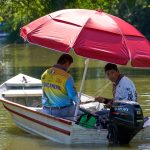 Dois homens pescam em um barco a motor com um guarda-sol vermelho sobre um rio tranquilo.
