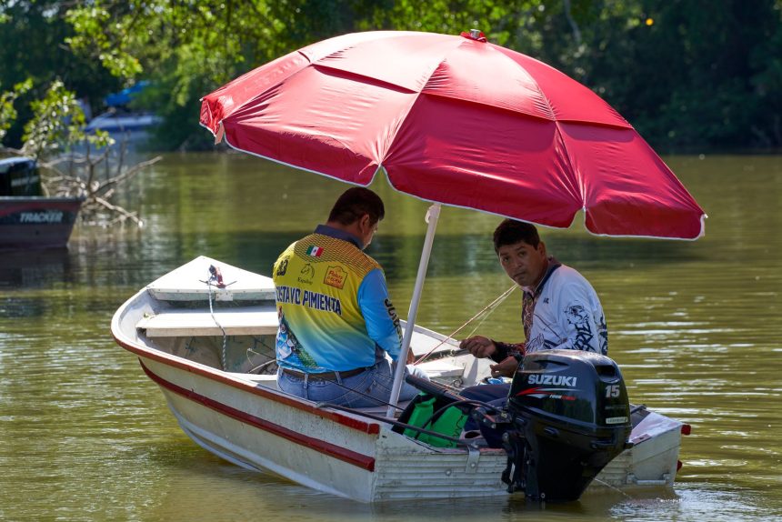 Dois homens pescam em um barco a motor com um guarda-sol vermelho sobre um rio tranquilo.